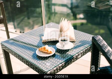 Tasse schwarzen Kaffee, frisch gebackenes Croissant und offenes Buch auf Rattantisch auf dem Hotelbalkon vor verschwommener grüner Resortlandschaft. Morgen bre Stockfoto