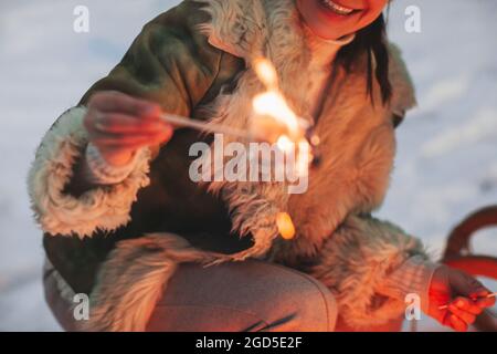 Eine glückliche Frau, die im Winterwald über dem Lagerfeuer süßes Moorgelchen röstet, und eine lächelnde Frau, die im Sitzen Essen zubereitete, aus der Nähe Stockfoto