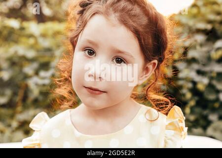 Nahaufnahme Porträt von niedlichen kleinen entzückenden Rotschopf Mädchen mit lockigen Haaren mit Zopf tragen gelb gepunkteten Kleid Blick auf Kamera beim Spielen in der Natur Stockfoto