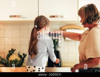 Liebevolle Großmutter kocht zusammen mit Enkelin zu Hause, verwendet Mixer, um Gemüse zu mischen, kleines Mädchen lernt, wie man Geräte mit Grand zu verwenden Stockfoto