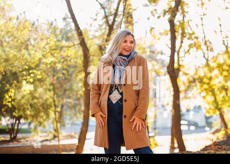 Eine glückliche Frau mittleren Alters genießt den Herbst draußen, in legerer Kleidung und beigem Mantel, der im gelben Park steht, zur Seite schaut und fröhlich lächelt Stockfoto