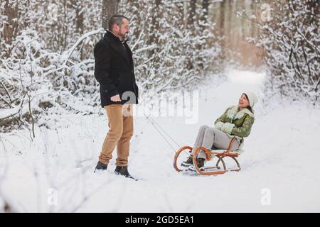 Glücklich verspielt reifes Familienpaar Rodeln im Winterpark, lachen und Spaß zusammen haben, positive Frau mittleren Alters sitzt auf Schlitten, während ihre Hus Stockfoto