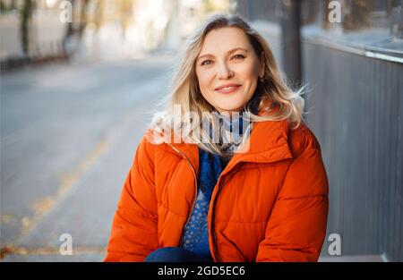 Glücklich lächelnde Frau mittleren Alters in orangefarbener Daunenjacke sitzt auf Betontreppen im Freien und blickt mit positivem Gesichtsausdruck auf die Kamera, ruht sich aus Stockfoto