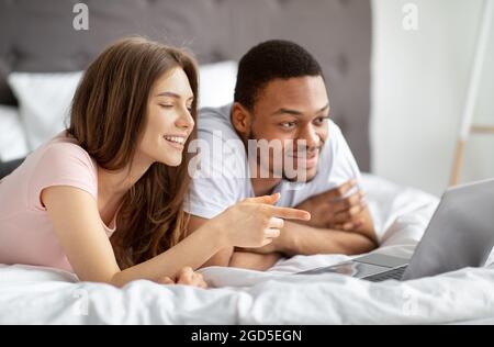 Fröhliches, tausendjähriges multinationales Paar, das mit einem Laptop auf dem Bett liegt und sich gemeinsam ein unterhaltsames Video ansieht. Glücklicher schwarzer Kerl und kaukasische Frau im Internet surfen, Stockfoto