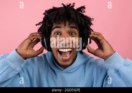 Überglücklich schwarzer Teenager, der beliebte Musik im Headset genießt und den Mund vor Aufregung auf rosafarbenem Studiohintergrund öffnet. Schöner Afro teen liebevoller cooler Song Stockfoto
