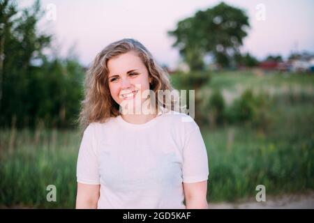 Portrait Of Beautiful Plus Size Junge Frau In Weißem Shirt Posiert Im Sommer Feld Wiese Bei Sonnenuntergang Hintergrund Stockfoto