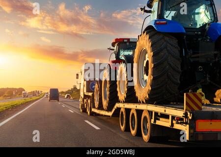 POV schweren Flurförderzeug Sattelauflieger Flachbett Plattform Transport zwei große moderne Landwirtschaft Traktor Maschine auf gemeinsame Autobahn Straße bei Sonnenuntergang Sonnenaufgang Himmel Stockfoto