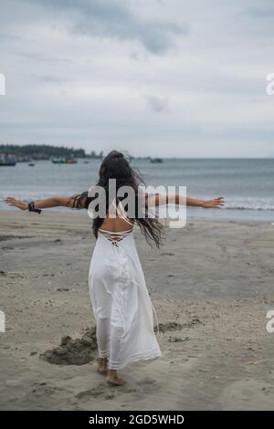 Schöne Frau in weißem Kleid zu Fuß am Strand. Hände zur Seite. Rückansicht. Meereshintergrund. Schwarzes langes lockiges Haar. Wind in ihren Haaren. Stockfoto
