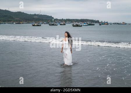 Charmante junge mongolische Frau in weißem Kleid zu Fuß am Strand. Schwarzes langes lockiges Haar. Blick beiseite. Romantisches Foto. Meerblick. Hochwertige Fotos Stockfoto