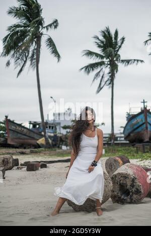 Friedliche junge asiatische Frau in weißem Kleid, die auf dem Stein sitzt und die Kamera mit Kopierraum anschaut. Brünett lange lockiges Haar. Romantisch. Stockfoto