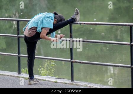 MULTITASKING. Eine Frau streckt ihren Oberschenkel und liest gleichzeitig ihr Handy. Im Kissena Park in Flushing, Queens, New York City. Stockfoto