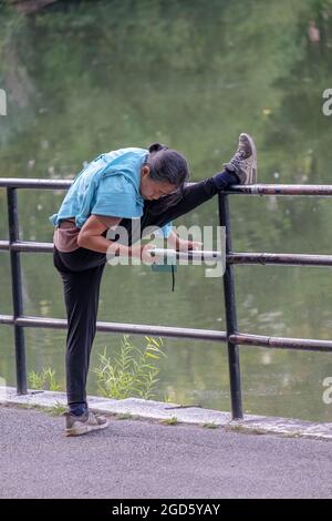 MULTITASKING. Eine Frau streckt ihren Oberschenkel und liest gleichzeitig ihr Handy. Im Kissena Park in Flushing, Queens, New York City. Stockfoto