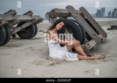 Schöne junge asiatische Frau in weißem Kleid sitzt am Strand und hält ihr Bein. Schwarzes langes lockiges Haar. Romantisches Foto. Hochwertige Fotos Stockfoto