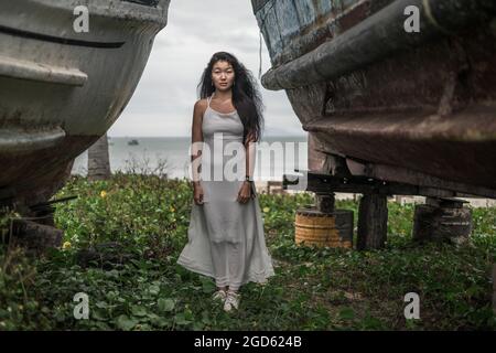 Charmante junge mongolische Frau in hellgrauem Kleid, die zwischen zwei alten Holzschiffen steht. Schwarzes langes lockiges Haar. Kamera mit Kopierplatz betrachten. Stockfoto