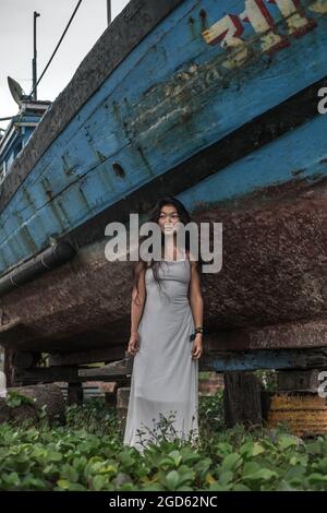 Schöne junge mongolische Frau in weißem Kleid, die in der Nähe des alten Holzschiffs steht. Brünett lange lockiges Haar. Blick beiseite. Romantisches Foto. Hochwertige Fotos Stockfoto