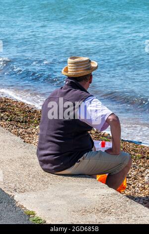 Mann, der auf einer Meereswand sitzt und auf das Meer blickt, Mann mit Hut am Strand, Mann mit Sommerhut, Mann, der an der Strandwand sitzt und denkt. Stockfoto