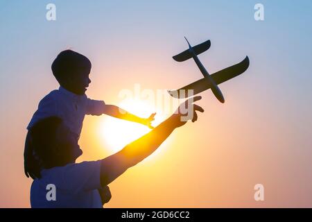 Die Silhouette eines Jungen, der auf der Schulter seines Vaters sitzt, lässt vor dem Hintergrund der untergehenden Sonne ein Modellflugzeug in den Abendhimmel fliegen. Kinderz Stockfoto