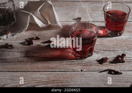 Heißer Hibiskustee in Gläsern auf Holztisch, Rosenblätter und trockener Tee auf weißem rustikalem Holztisch, Dampf steigt aus dem Glas Stockfoto