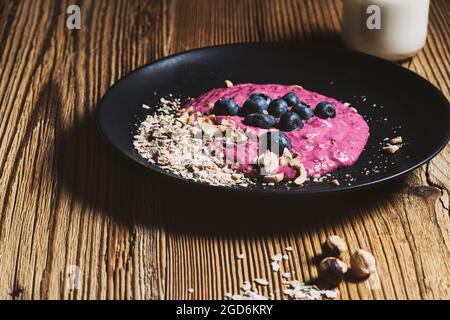 Heidelbeer-Smoothie-Schale zum Frühstück. Frische Beeren, gerollter Hafer, geröstete Haselnuss auf schwarzem Teller und Kefir im Glas Stockfoto