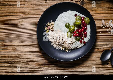 Frühstückssmoothie Schüssel mit Stachelbeeren, roten Johannisbeeren. Frische Beeren, Haferflocken, Pistazien auf schwarzem Teller über rustikalem Holztisch, Draufsicht Stockfoto