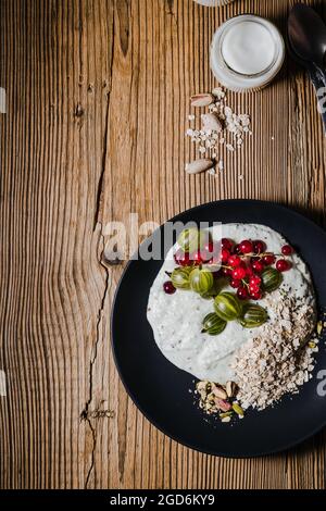 Frühstückssmoothie Schüssel mit Stachelbeeren, roten Johannisbeeren. Frische Beeren, gerollter Hafer, Pistazien auf schwarzem Teller und Krug Kefir über rustikalem Holztab Stockfoto