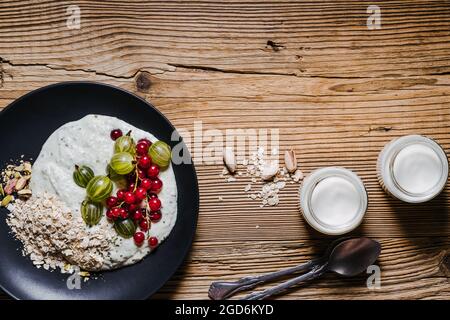 Frühstückssmoothie Schüssel mit Stachelbeeren, roten Johannisbeeren. Frische Beeren, gerollter Hafer, Pistazien auf schwarzem Teller und Krug Kefir über rustikalem Holztab Stockfoto