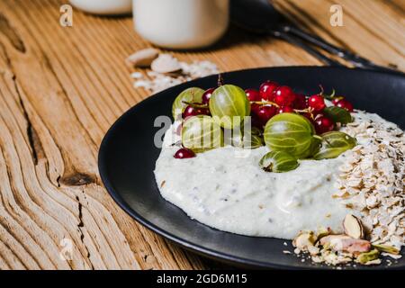 Frühstückssmoothie Schüssel mit Stachelbeeren, roten Johannisbeeren. Frische Beeren, gerollter Hafer, Pistazien auf schwarzem Teller und Krug Kefir über rustikalem Holztab Stockfoto