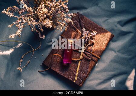 Überkopfbild einer Glasstopfflasche mit rosa, flüssigem, getrocknetem Lavendel auf einem ledergebundenen Buch auf einer grauen Oberfläche Stockfoto