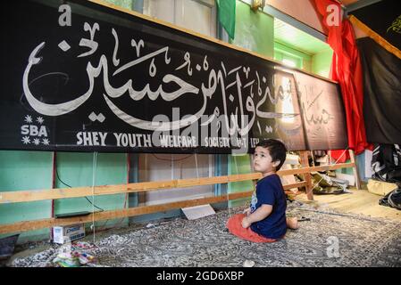 Srinagar, Indien. August 2021. Ein Kind sitzt vor einem Banner, das von einem Künstler vor Ashura entworfen wurde. Muharram ist der erste Monat des Islam. Es ist einer der heiligsten Monate im islamischen Kalender. Schiitische Muslime gedenken Muharram als einen Monat der Trauer zum Gedenken an das Martyrium des Enkels des islamischen Propheten Muhammad Imam Hussain, der 680 n. Chr. in der Schlacht von Karbala an der Ashura (10. Tag von Muharram) getötet wurde (Foto: Idrees Abbas/SOPA Images/Sipa USA) Quelle: SIPA USA/Alamy Live News Stockfoto