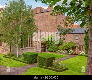 Ein kleiner Garten neben einem mittelalterlichen Gebäude. Eine Bank ist an einer alten Wand und vor einer Wiese aufgestellt. Ein weißer Baum und Hecken stehen im Vordergrund. Stockfoto