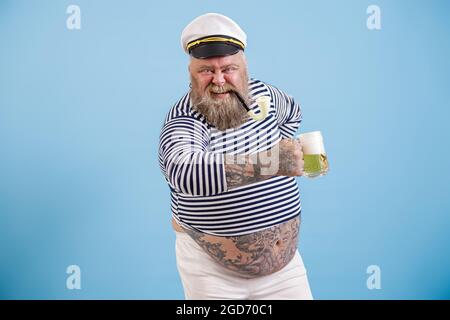 Ein glücklicher Mann in Übergröße im Matrosenanzug hält auf hellblauem Hintergrund köstliches Bier Stockfoto