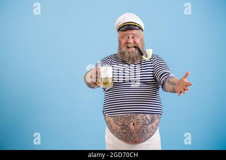 Fröhlicher übergewichtiger Seemann mit Rauchpfeife hält ein Glas frisches Bier auf hellblauem Hintergrund Stockfoto