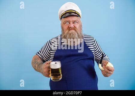 Lustige übergewichtige Seemann mit Schürze hält Rohr und leckeres Bier auf hellblauem Hintergrund Stockfoto