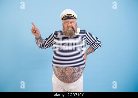 Plus Größe Seemann Mann mit Tattoos und rauchenden Pfeife zeigt neben Finger auf hellblauem Hintergrund Stockfoto