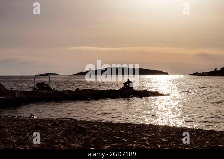 Silhouette eines Mannes, der bei Sonnenuntergang auf dem steinernen Pier sitzt und das Meer und entfernte Inseln beobachtet, fotografiert am Strand der Stadt Rogoznica, Kroatien Stockfoto