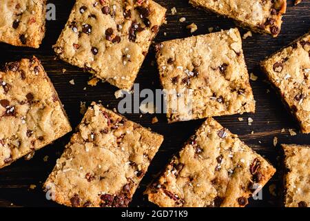 Brown Butter Bourbon Pecan Chocolate Chunk Blondies garniert mit flockigem Meersalz: Blondies in Quadrate geschnitten und auf einem dunklen Holzschneidebrett serviert Stockfoto