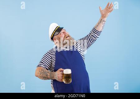 Positiv plump Mann in Seemann Kostüm hält Bier heben Hand auf blauem Hintergrund Stockfoto