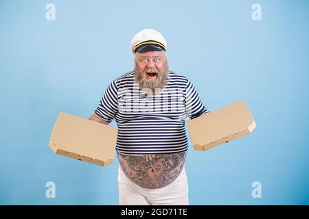 Aufgeregter Mann mit Übergewicht im Matrosenkostüm hält auf hellblauem Hintergrund Pizzaschachteln Stockfoto