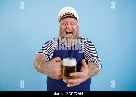 Ein fröhlicher, fetter Mann im Matrosenanzug bietet einen Becher frisches Bier auf hellblauem Hintergrund an Stockfoto