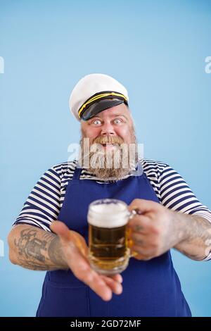 Fröhlicher, praller Mann im Matrosenkostüm bietet einen Glas Becher Bier auf hellblauem Hintergrund an Stockfoto