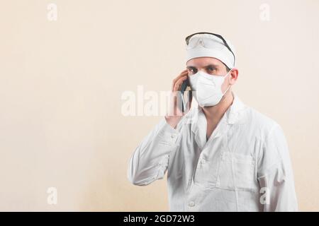 Junger Arzt in weißem Laborkittel, Schutzbrille und schützender Atemmaske, die das Mobiltelefon isoliert hält, um mit dem Handy zu sprechen. Stockfoto