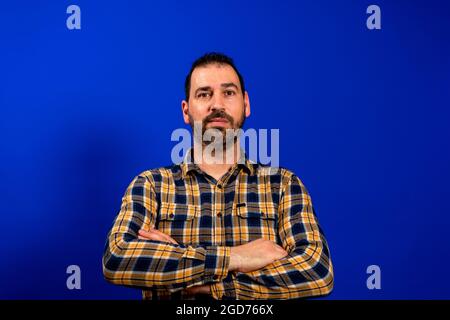 Der junge Mann, der über gekreuzte Arme steht, wirkt genervt über die Gefühle des Stirnrunzens auf blauem Hintergrund. Stockfoto