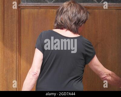 Rückseite einer Frau mit einem grauen T-Shirt vor einer alten braunen Tür, die sich bald öffnen wird Stockfoto