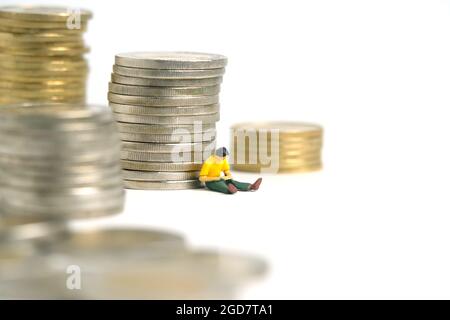 Miniatur Menschen Spielzeug Figur Fotografie. Ein männlichem Student, der vor einem Geldmünzstapel sitzt, isoliert auf weißem Hintergrund. Bildfoto Stockfoto