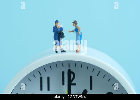 Miniatur Menschen Spielzeug konzeptuelle Fotografie. Termingerecht geliefert. Postboten, der über der Uhr stand, isoliert auf blauem Hintergrund. Stockfoto
