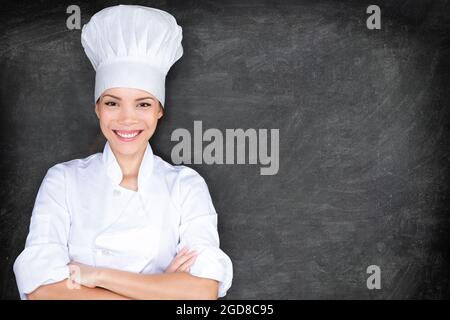 Asiatische Chefin Frau glücklich Porträt auf schwarzem Hintergrund Tafel Textur mit Copy Space. Junge professionelle Köchin bei der Arbeit in weißer Uniform und Haube Stockfoto