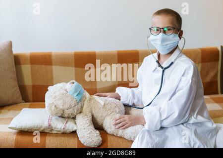 Blick auf den Kinderarzt oder die Krankenschwester mit Teddybär im Sonnenlicht zu Hause. Happy Boy hört einem Stethoskop-Spielzeug zu. Rollenspiel verspielter Kerl. In einer Brille und einem Gewand mit Schutzmaske Stockfoto