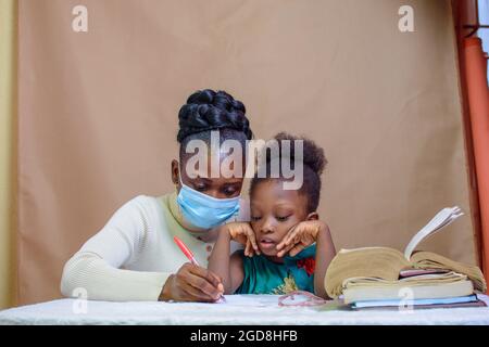 Afrikanische Lehrerin oder Mutter mit Nasenmaske, die einem kleinen Mädchen beim Studium beibringt und hilft, sich in Schule, Bildung und Karriere zu übertreffen Stockfoto