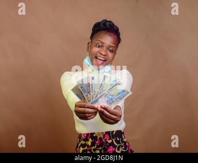 Fröhliche, süße afrikanische Dame mit Nasenmaske, die mehrere Naira-Scheine, Bargeld, nigerianische Währung oder Geld für Transaktionen in ihren Händen hält Stockfoto