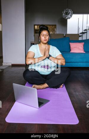 Junge kurvige lateinerin mit Computer auf Matte sitzend, meditierend in Mexiko, plus weibliche Größe Stockfoto